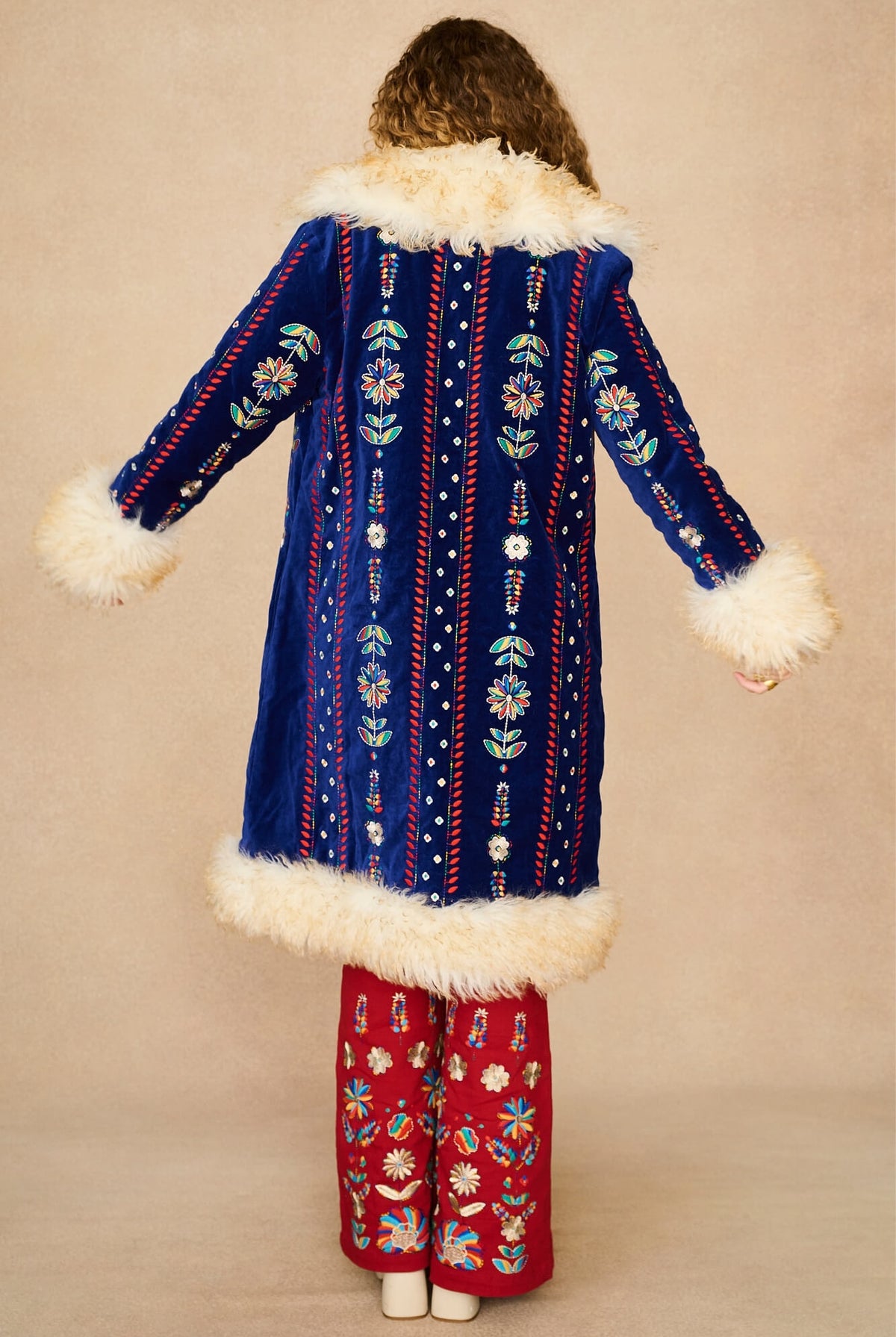 A model shown from the back wearing a long blue velvet Penny Lane coat with white faux-fur trim and colourful embroidered vertical patterns, paired with red patterned pants, against a plain beige studio background.