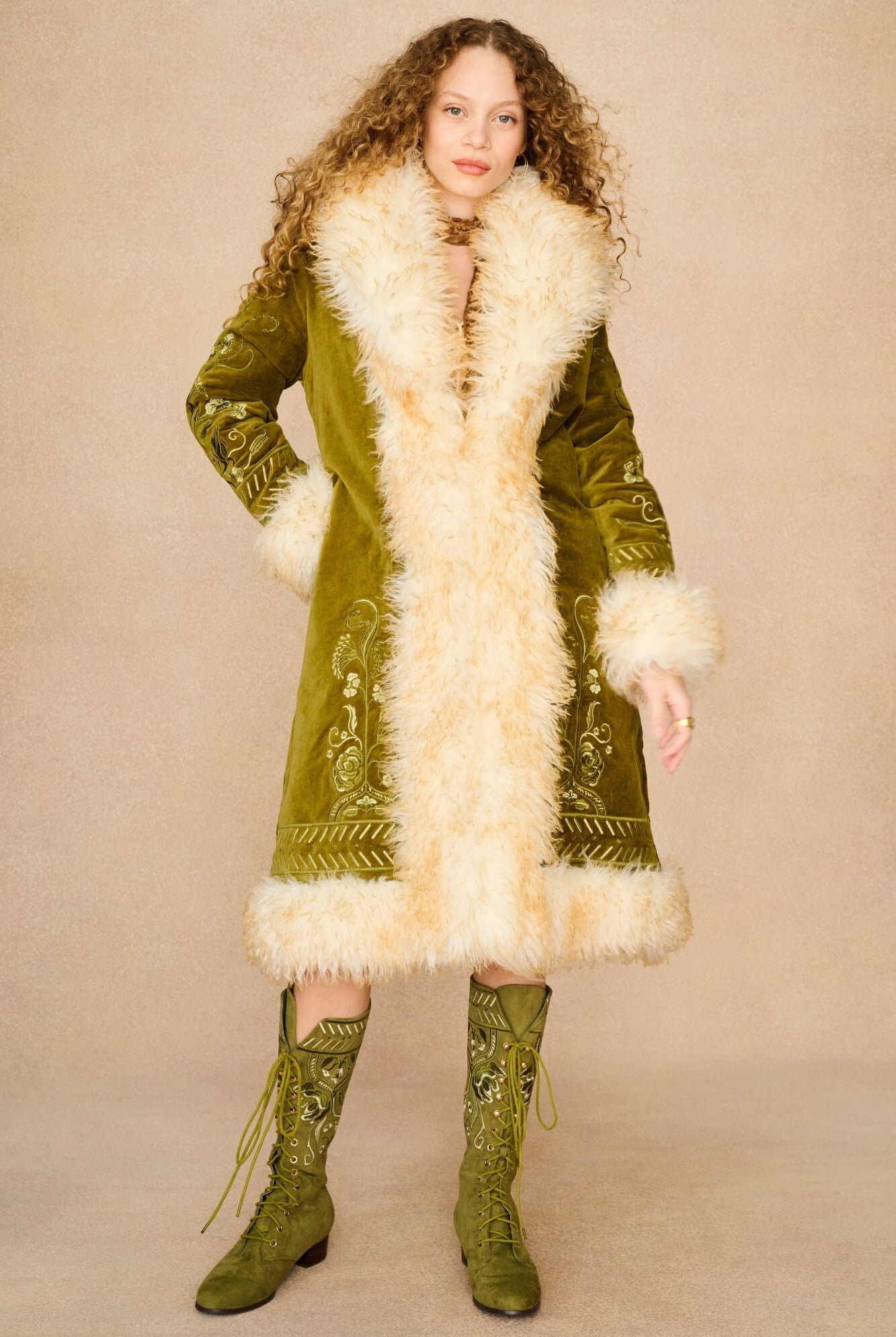 A model wearing a long olive green velvet Penny Lane coat by The Hippie Shake with a large cream faux-fur collar and hem, paired with matching green embroidered lace-up boots, against a solid beige studio background.