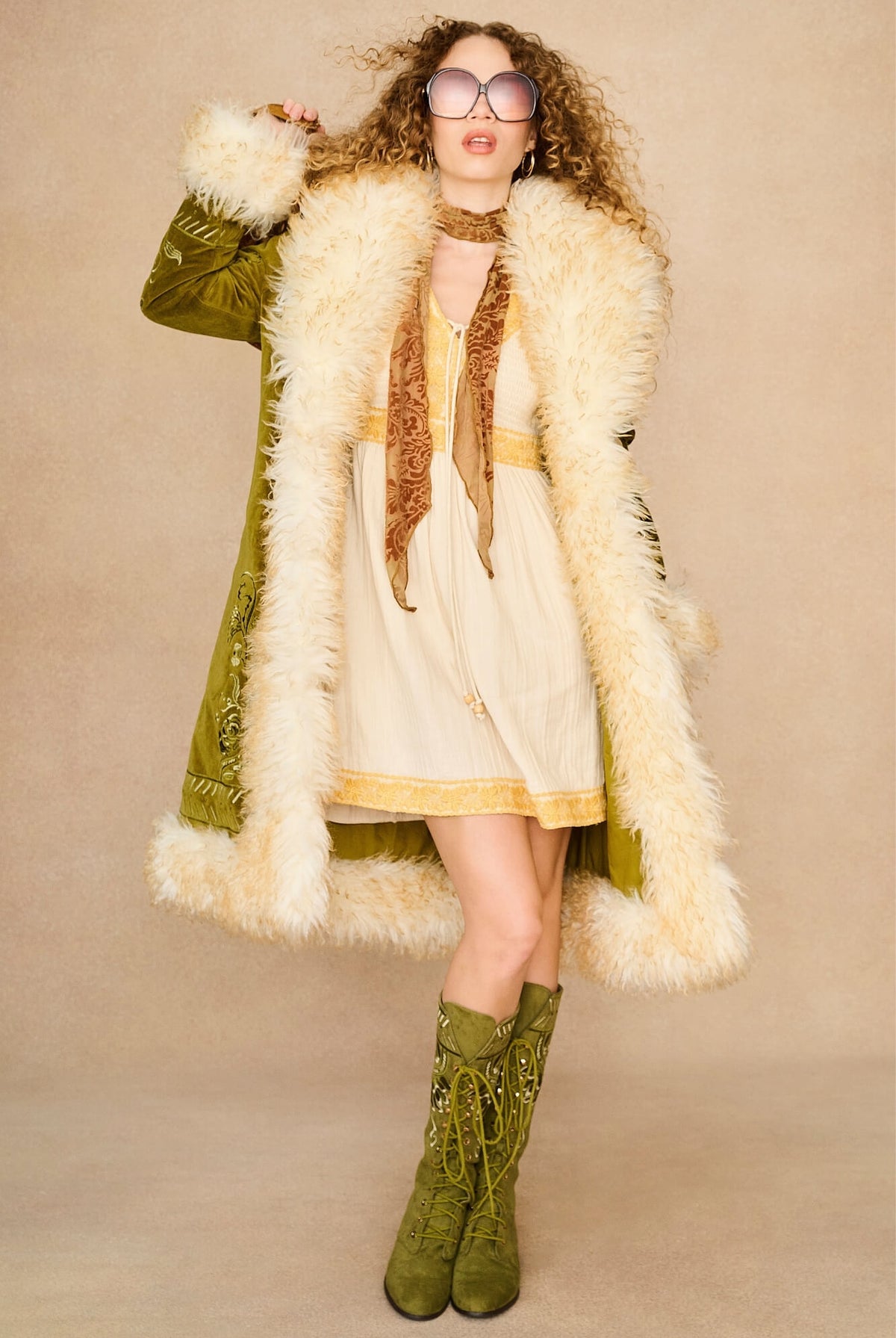 A woman modeling a bohemian-style outfit featuring an olive green embroidered Penny Lane coat with a fluffy cream faux-fur collar, a cream dress with gold trim, a brown patterned scarf, and matching olive green lace-up boots, set against a solid beige studio background.