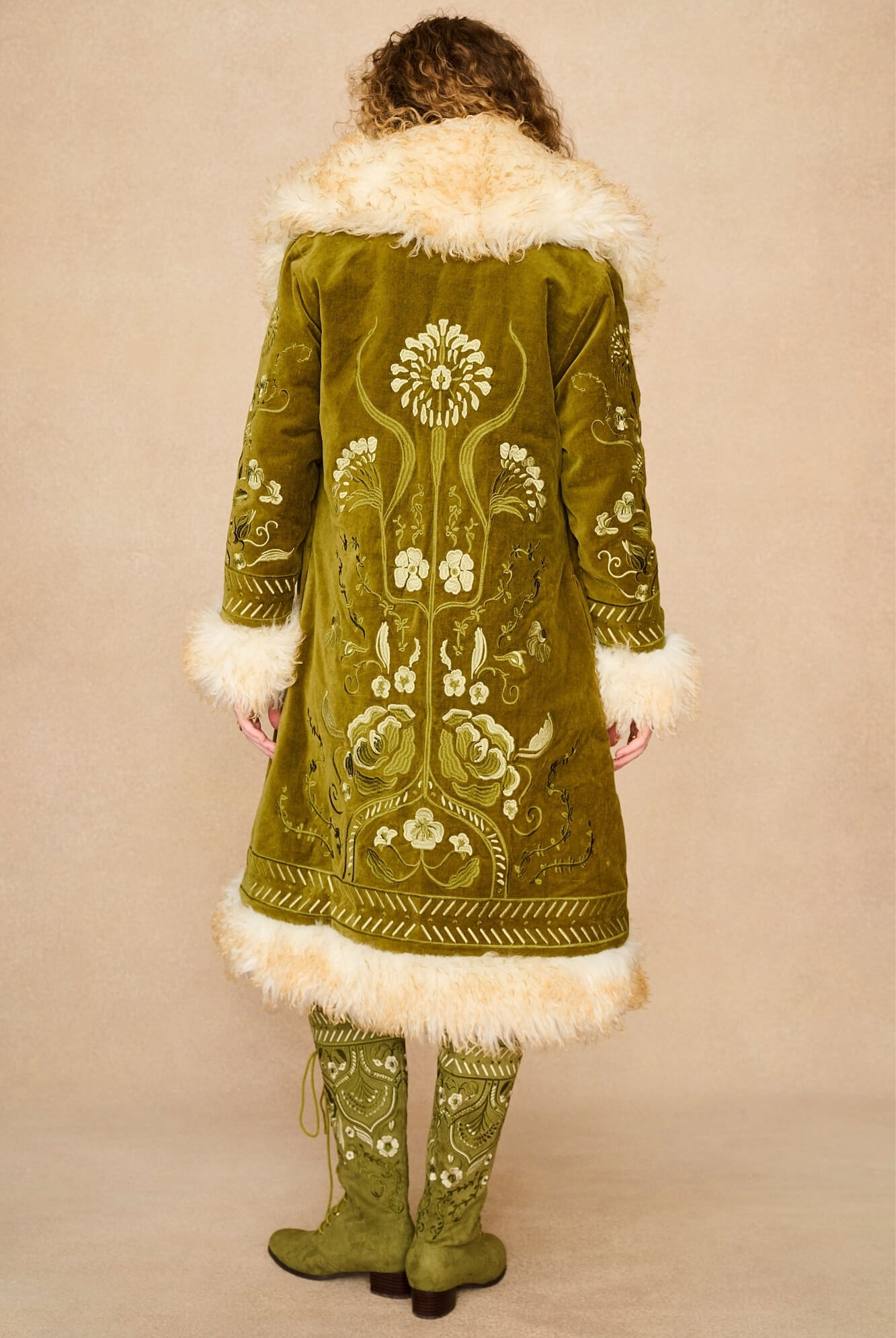 Model shown from the back wearing a long olive green embroidered Penny lane coat by The Hippie Shake with cream fur trim and matching embroidered green boots, against a plain beige studio background.