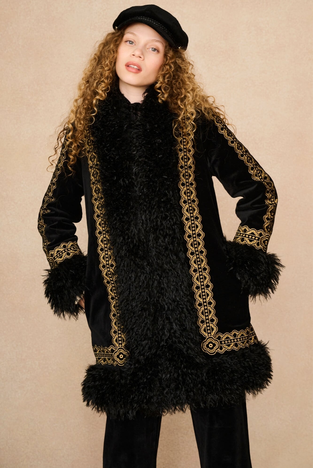 A model with long curly blonde hair wearing a black velvet and faux fur Penny Lane coat by The Hippie Shake with ornate gold embroidered borders, a black beret, and black pants, posed against a solid beige studio background.