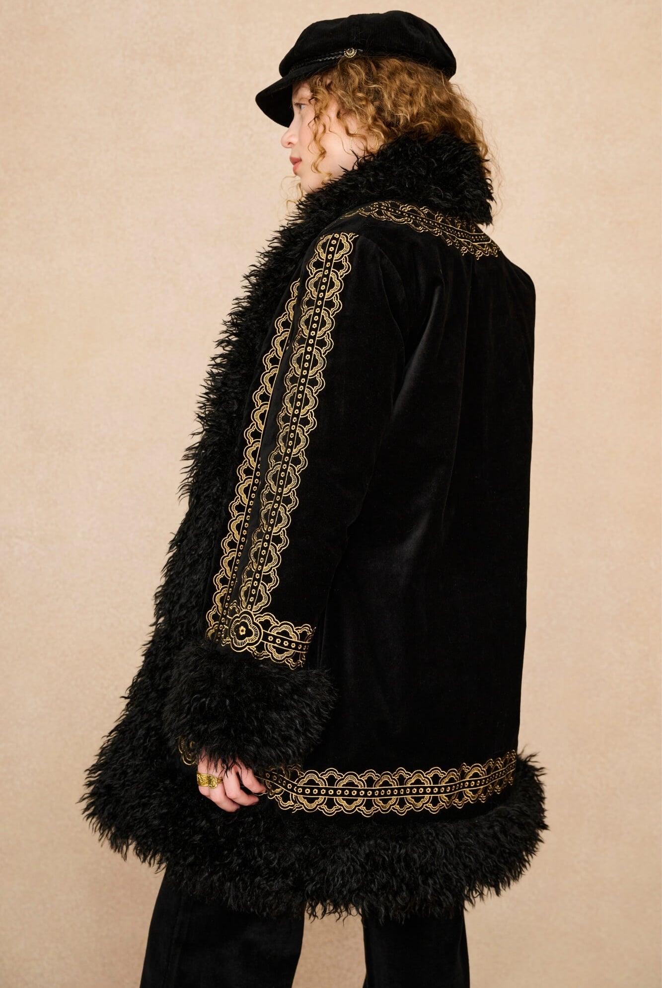 A person shown in side profile wearing a black velvet coat with ornate gold embroidered trim and a black feathered collar, along with a black cap, against a plain beige studio background.