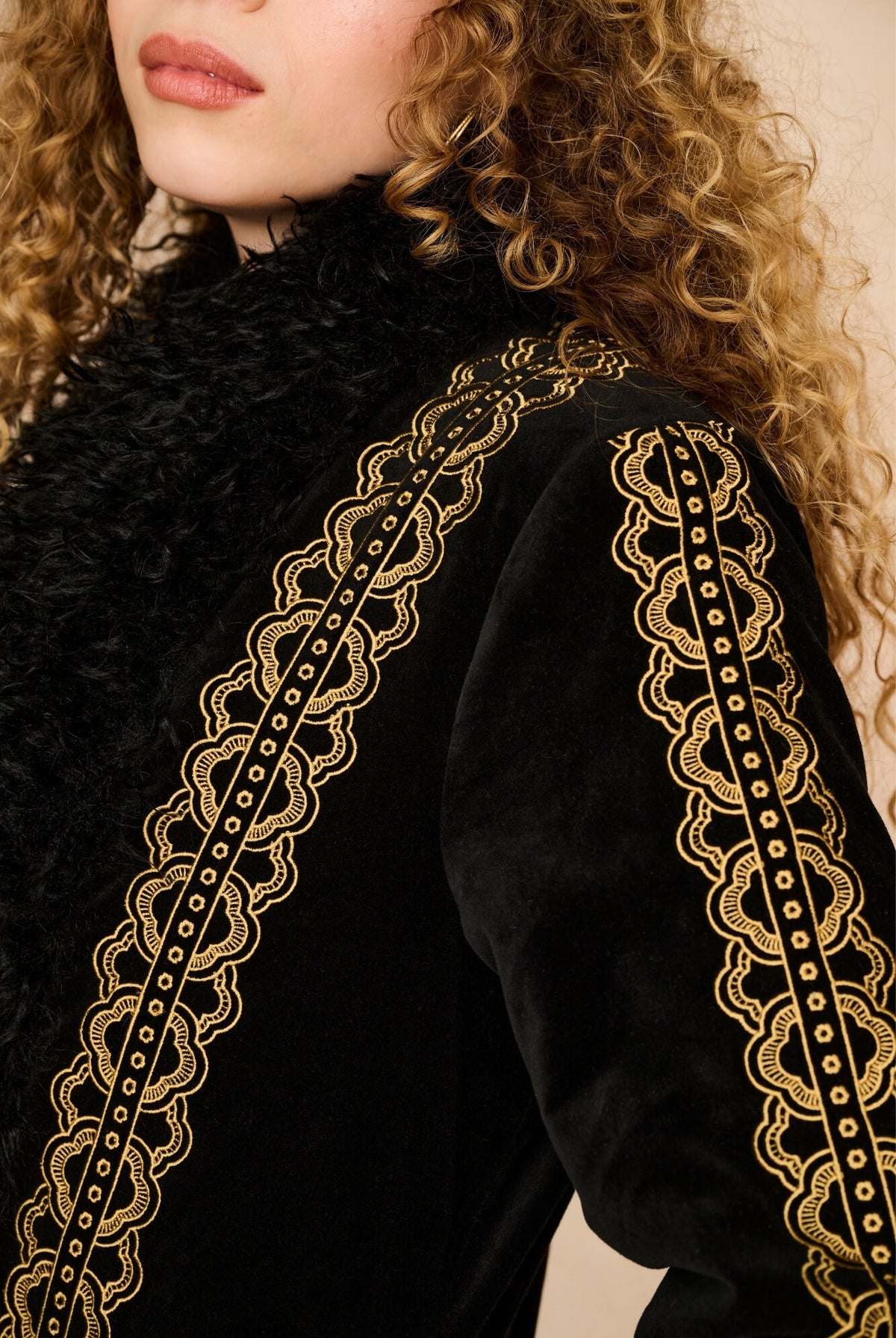 Close-up of a model wearing a black velvet jacket with intricate gold embroidered lace trim along the shoulders and collar.