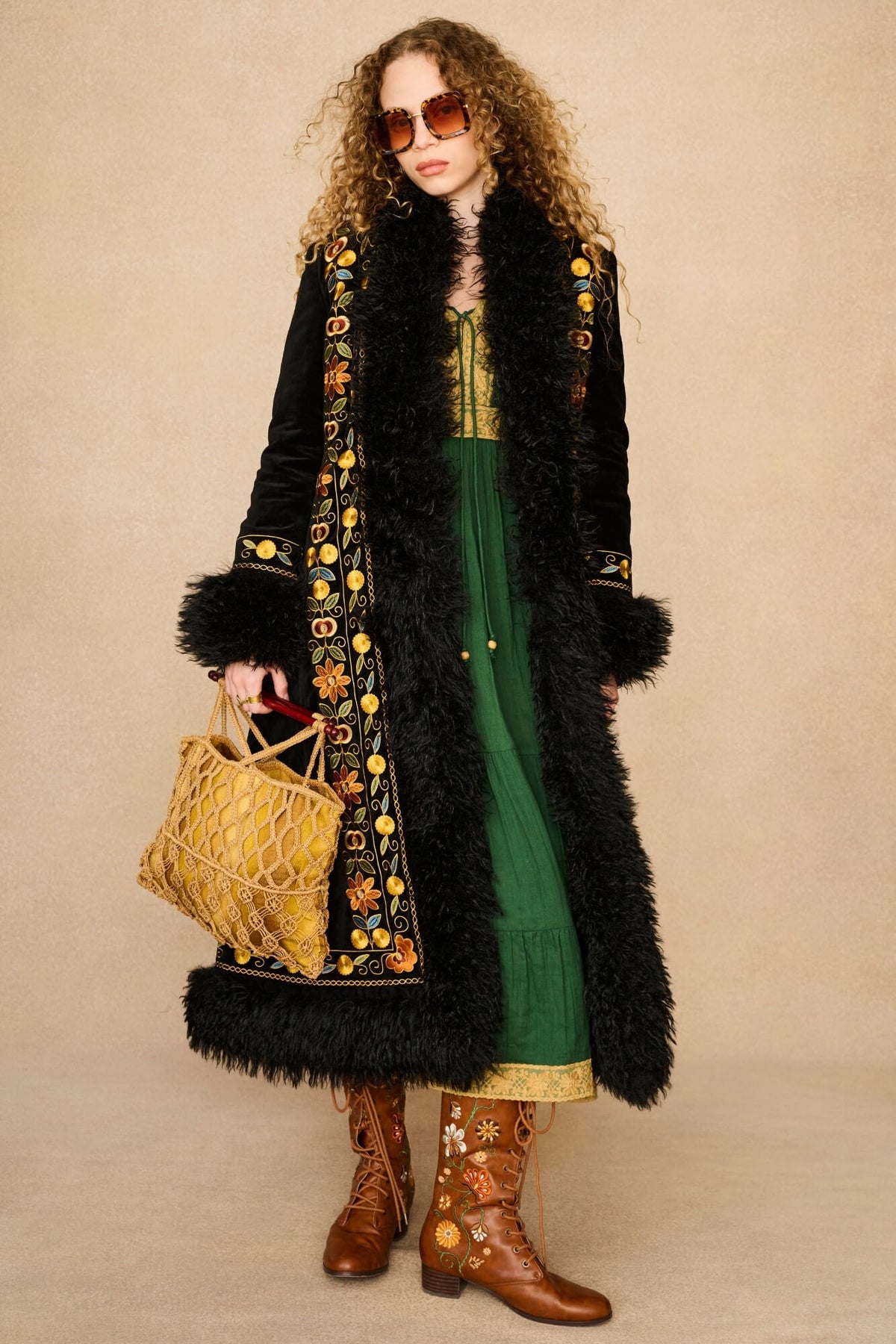 A woman stands against a plain beige studio background wearing a long black faux-fur coat with ornate gold floral embroidery, a green dress, brown western boots, and holding a woven tan tote bag.