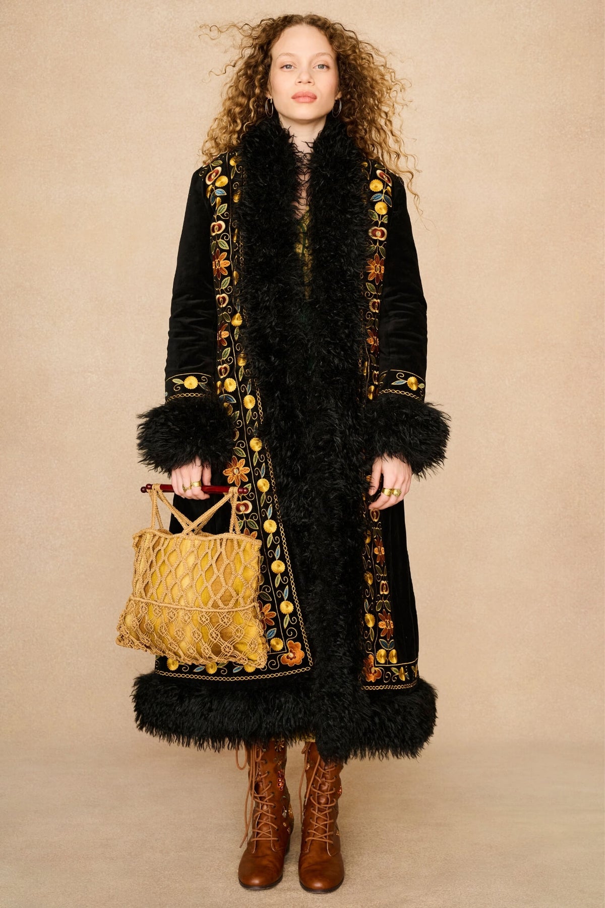 A model stands against a beige studio backdrop wearing a long black velvet Penny Lane Coat by The Hippie Shake with black faux-fur trim and ornate gold, yellow, and orange embroidered detailing along the front and cuffs. She is holding a yellow woven handbag and wearing brown lace-up ankle boots.
