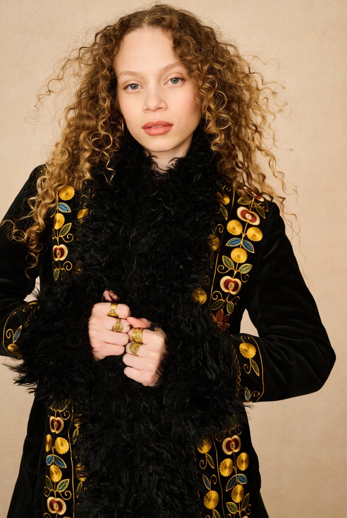 A model with curly light brown hair wearing a black velvet Penny Lane coat with gold floral embroidery and a fluffy black feather collar, posed against a neutral beige background.