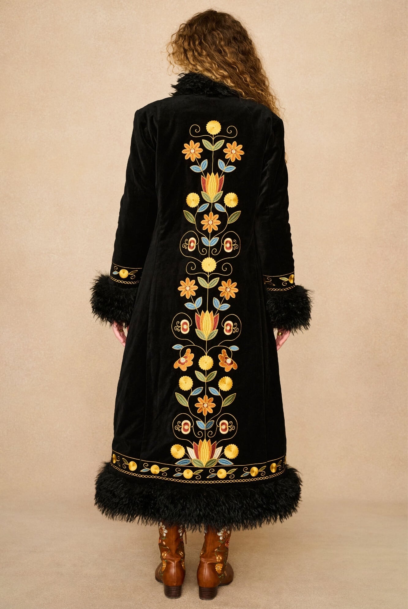 A woman viewed from behind wearing a long black velvet Penny lane coat by The Hippie Shake with colourful floral embroidery down the center, black feather trim at the collar and hem, and brown leather boots, against a plain beige studio background.