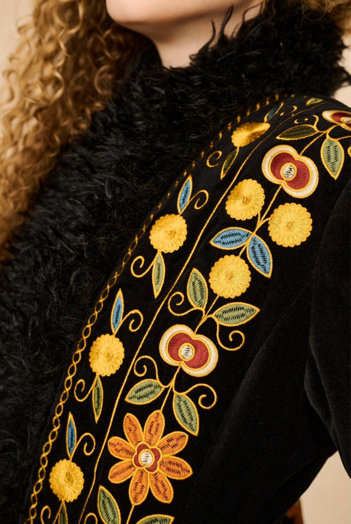 Close-up of a black velvet coat with a black faux-fur collar and ornate multicolor floral embroidery in yellow, gold, blue, and red along the neckline.
