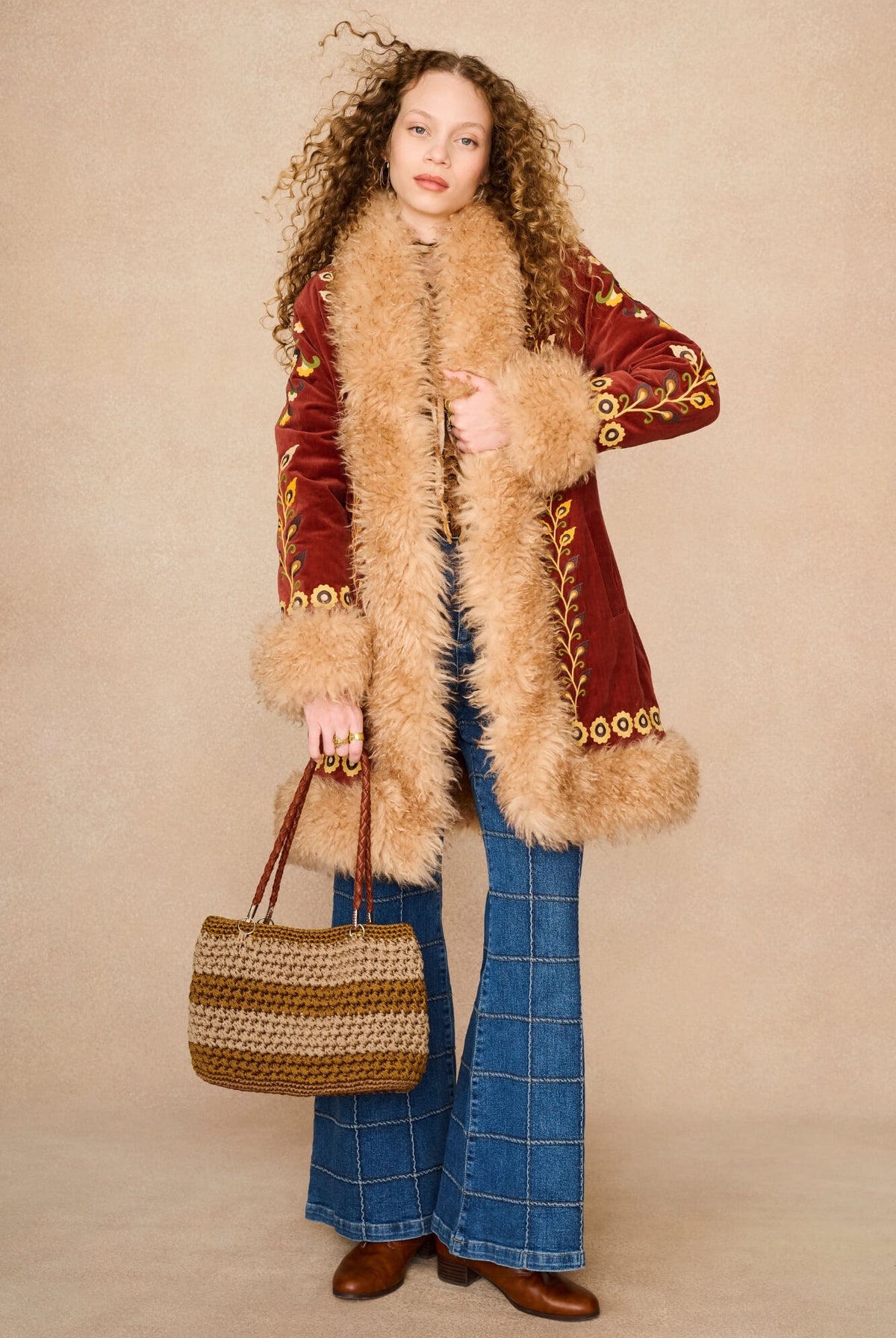 A model wearing a vintage-style red velvet Penny lane by The Hippie Shake coat with beige fur trim and gold embroidery, paired with blue plaid flared jeans, brown boots, and a woven tan handbag, set against a plain beige studio backdrop.