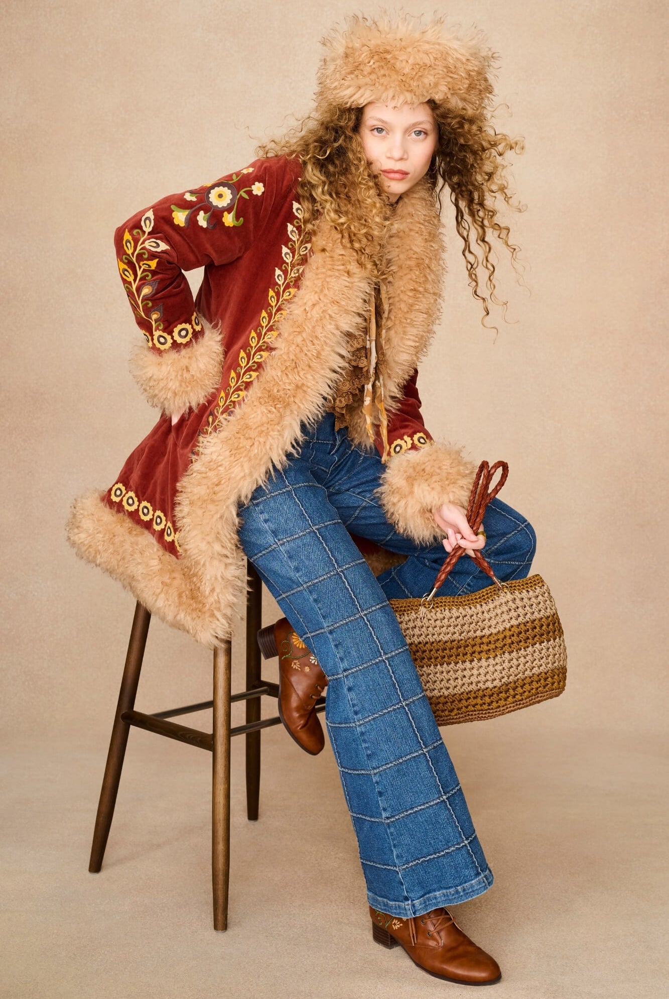 A model sits on a wooden stool wearing a vintage-style red velvet jacket with beige fur trim, a matching fur-trimmed hat, blue plaid jeans, brown leather boots, and holding a woven straw handbag against a beige studio background.