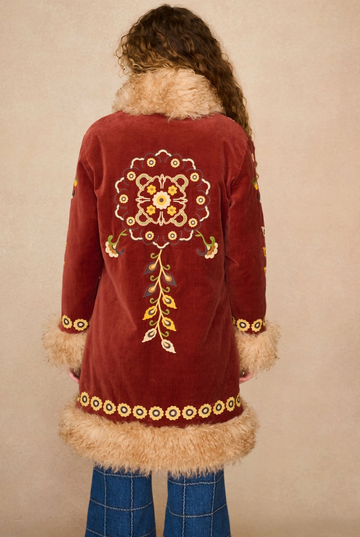 A person viewed from the back wearing a burgundy velvet Penny Lane Coat with beige fur trim and colourful embroidered floral patterns, paired with blue jeans, against a plain beige background.