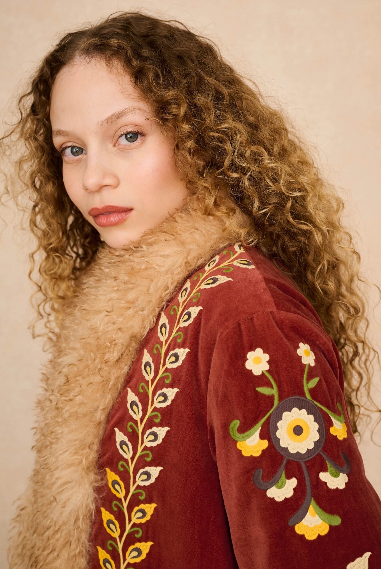 A close-up portrait of a woman with long curly brown hair wearing a burgundy velvet jacket with a beige fur collar and colorful floral embroidery on the sleeve, set against a plain beige background.