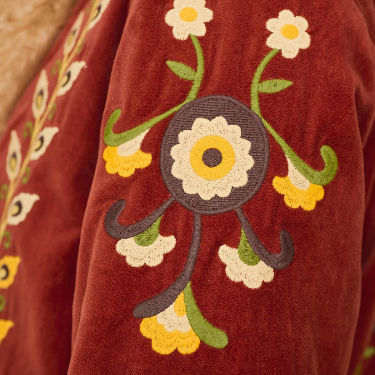 Close-up of a rust red velvet jacket with embroidered floral patterns on the sleeve, featuring white, yellow, and green details.