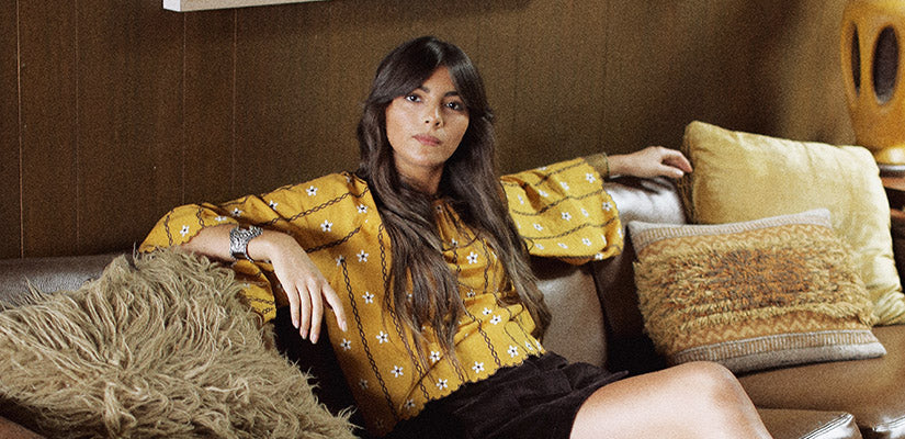 Hero banner for The Hippie Shake's boho and 70s blouses and tops - Model wears yellow embroidered smock top sat on sofa.