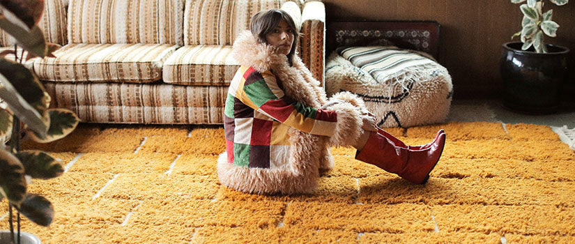 Hero banner for boho and 70s style coats and jackets from The Hippie Shake - Model wearing patchwork Penny Lane coat sat on yellow rug.