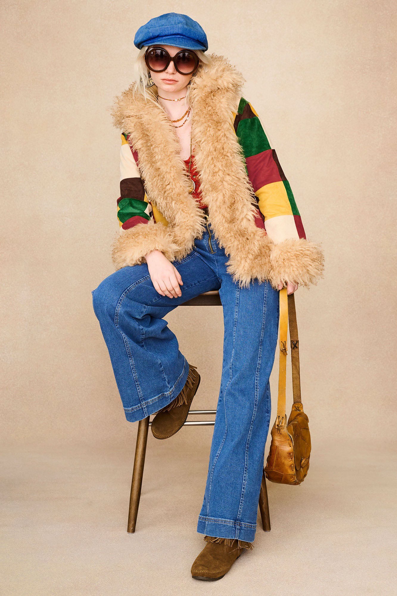 Model seated in The Hippie Shake's patchwork Penny Lane coat with fluffy trims and wide-leg jeans.