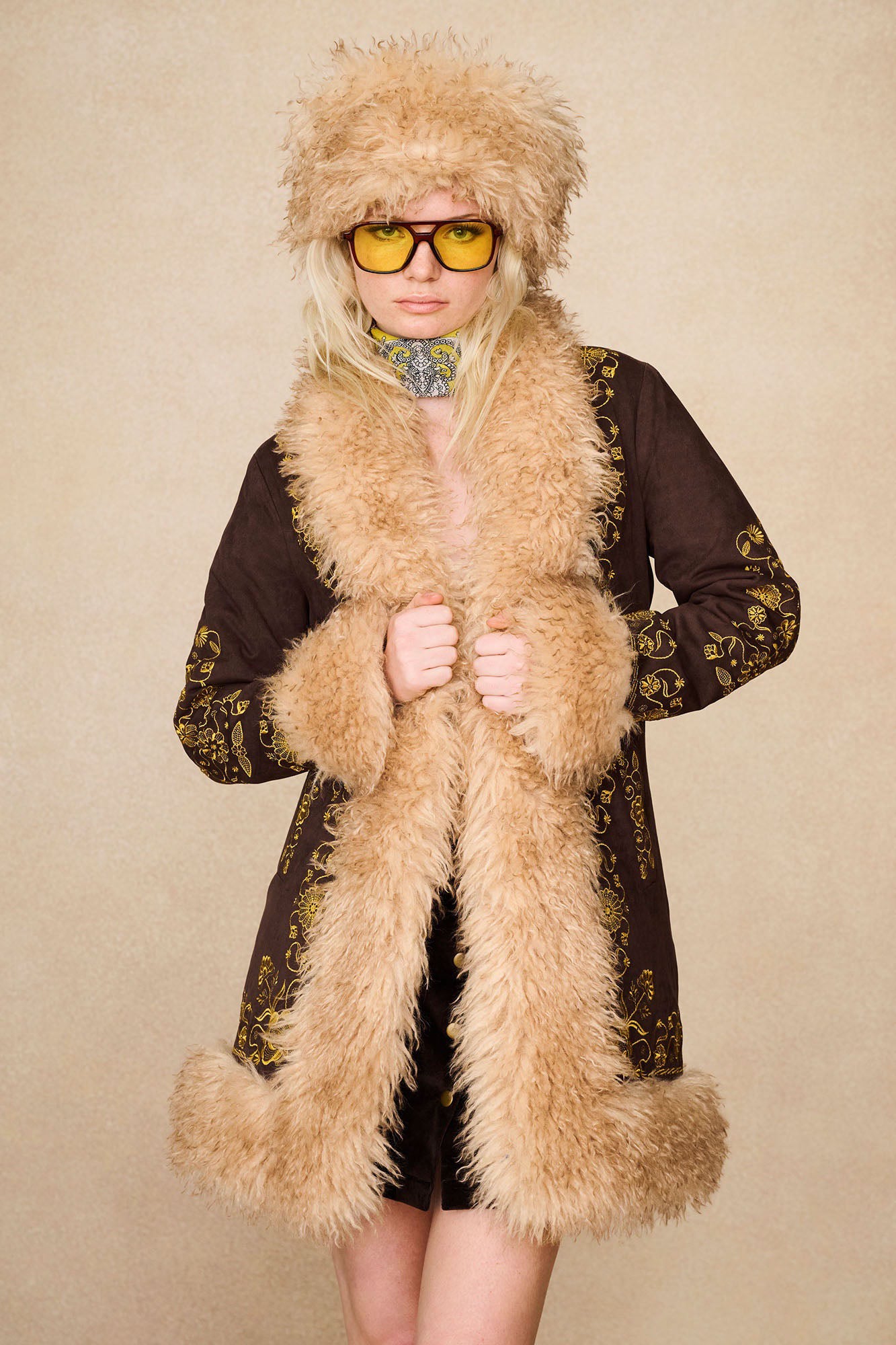 Model wearing The Hippie Shake's brown embroidered Penny Lane coat, styled with our brown faux fur hat and yellow sunglasses. 