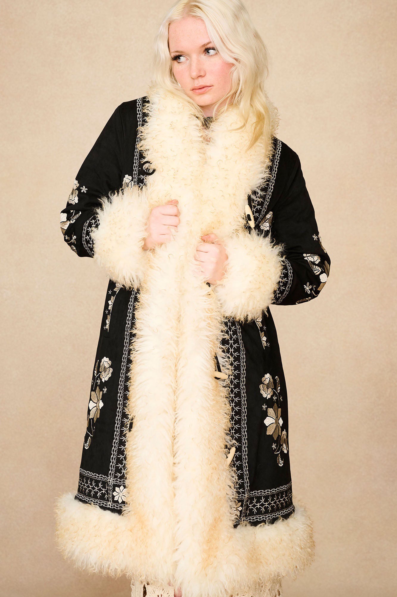 Model holding The Hippie Shake's black embroidered Penny Lane coat closed, highlighting the off-white shaggy faux fur trims.