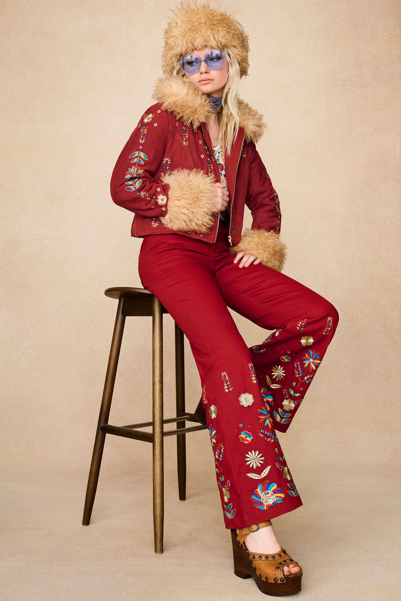 Model sitting on a stool wearing The Hippie Shake's red embroidered flared trousers with the matching jacket.