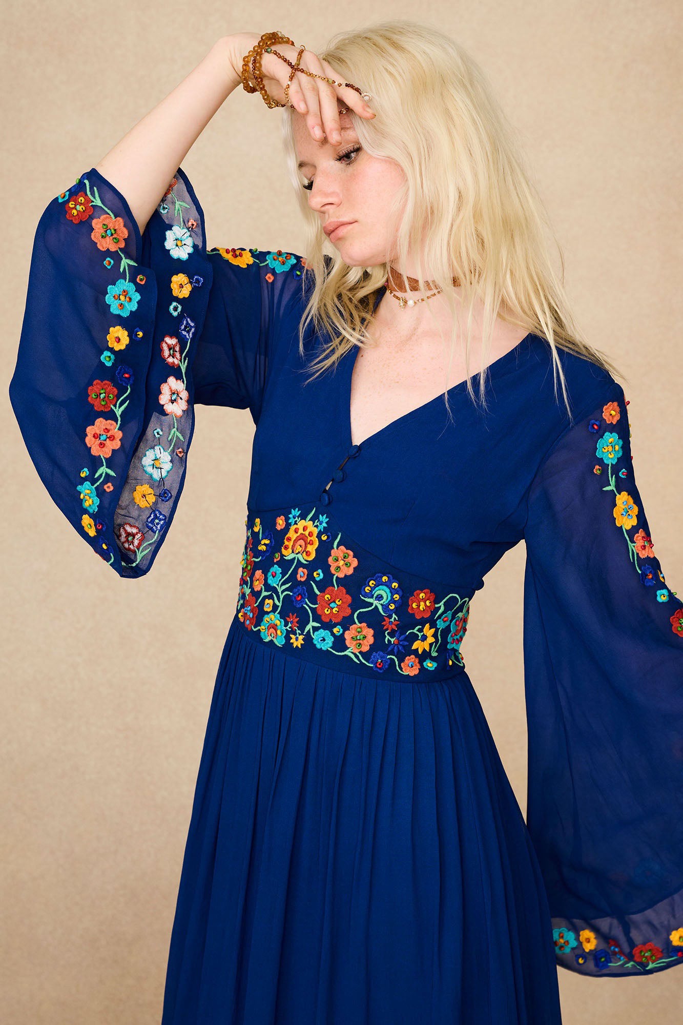 Model wearing The Hippie Shake's blue floral embroidered maxi dress with flared sleeves and hand resting on forehead.
