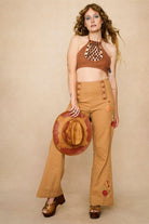 Woman wearing a 70s style brown halter top with embellishments and high-waisted pants against a beige background - outfit front view