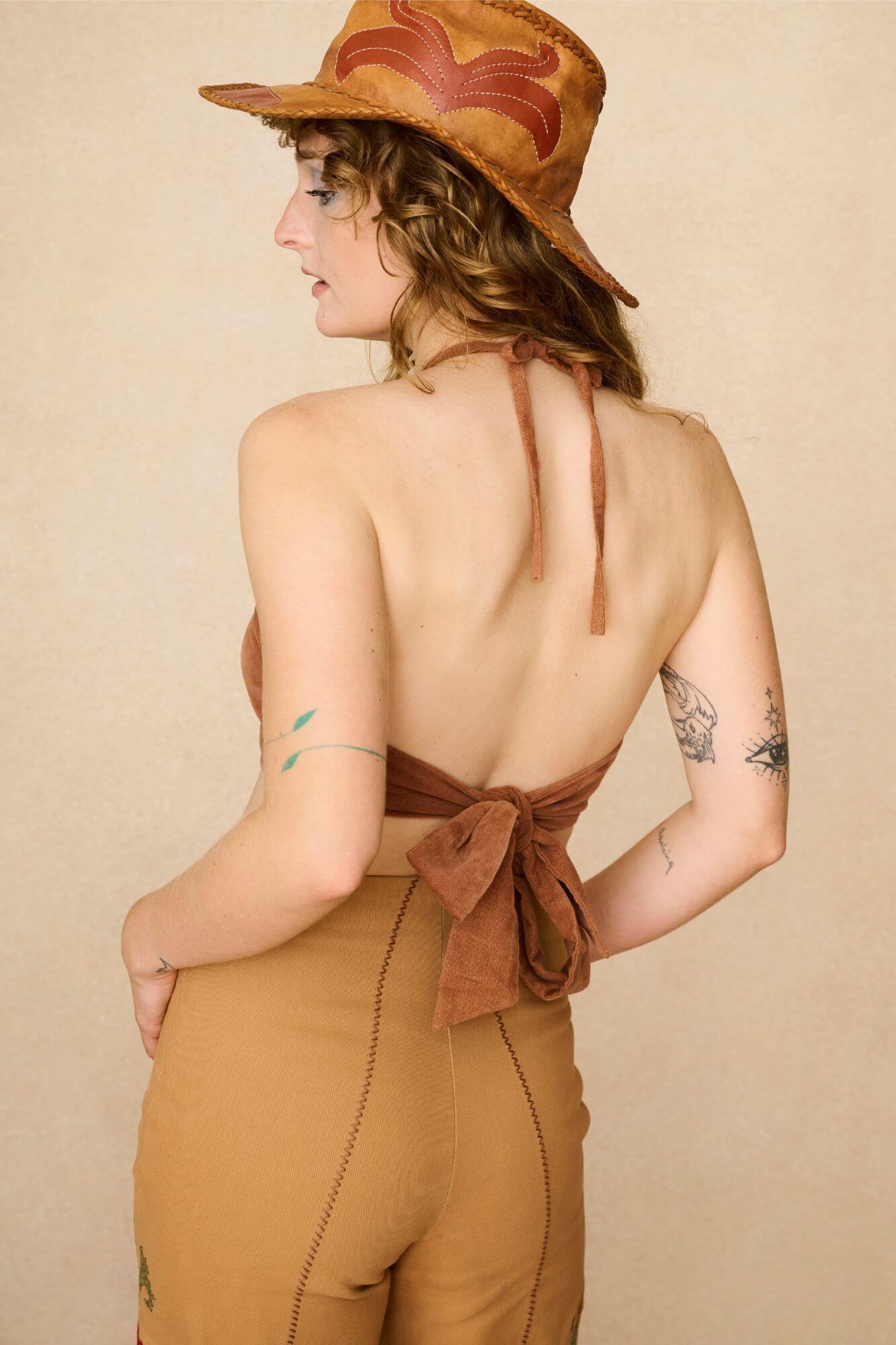 Woman wearing a 70s style brown halter top with embellishments and high-waisted pants against a beige background - back view