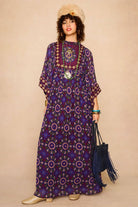 Model wears a 70s style bohemian maxi dress in a printed purple fabric with hand embroidery
