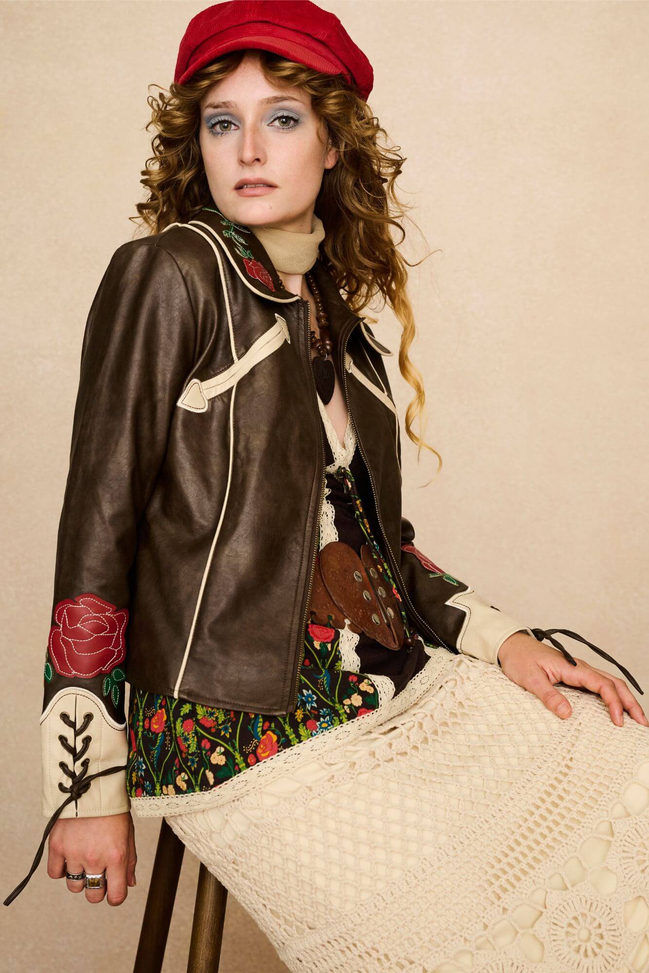 Model wearing a brown 70s style faux leather jacket with red floral embroidery against a beige background - seated outfit view