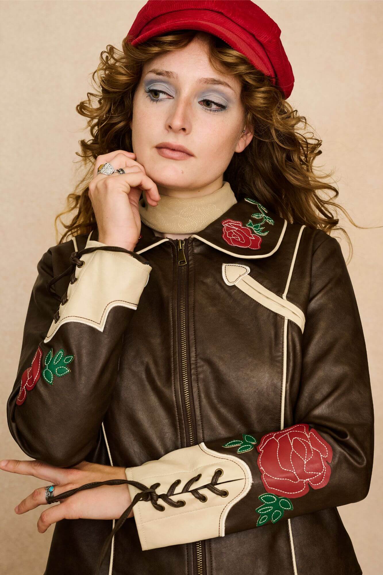 Model wearing a brown 70s style faux leather jacket with red floral embroidery against a beige background - close front view
