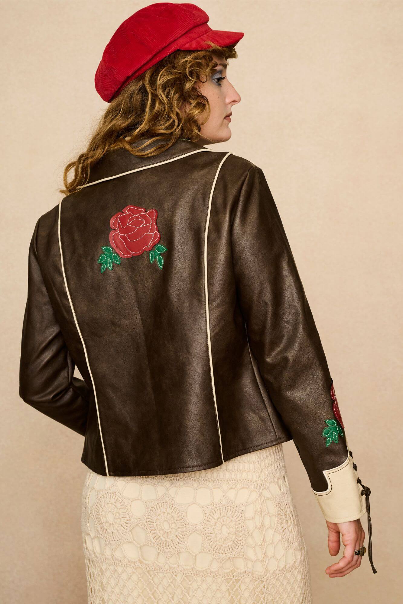 Model wearing a brown 70s style faux leather jacket with red floral embroidery against a beige background - back view