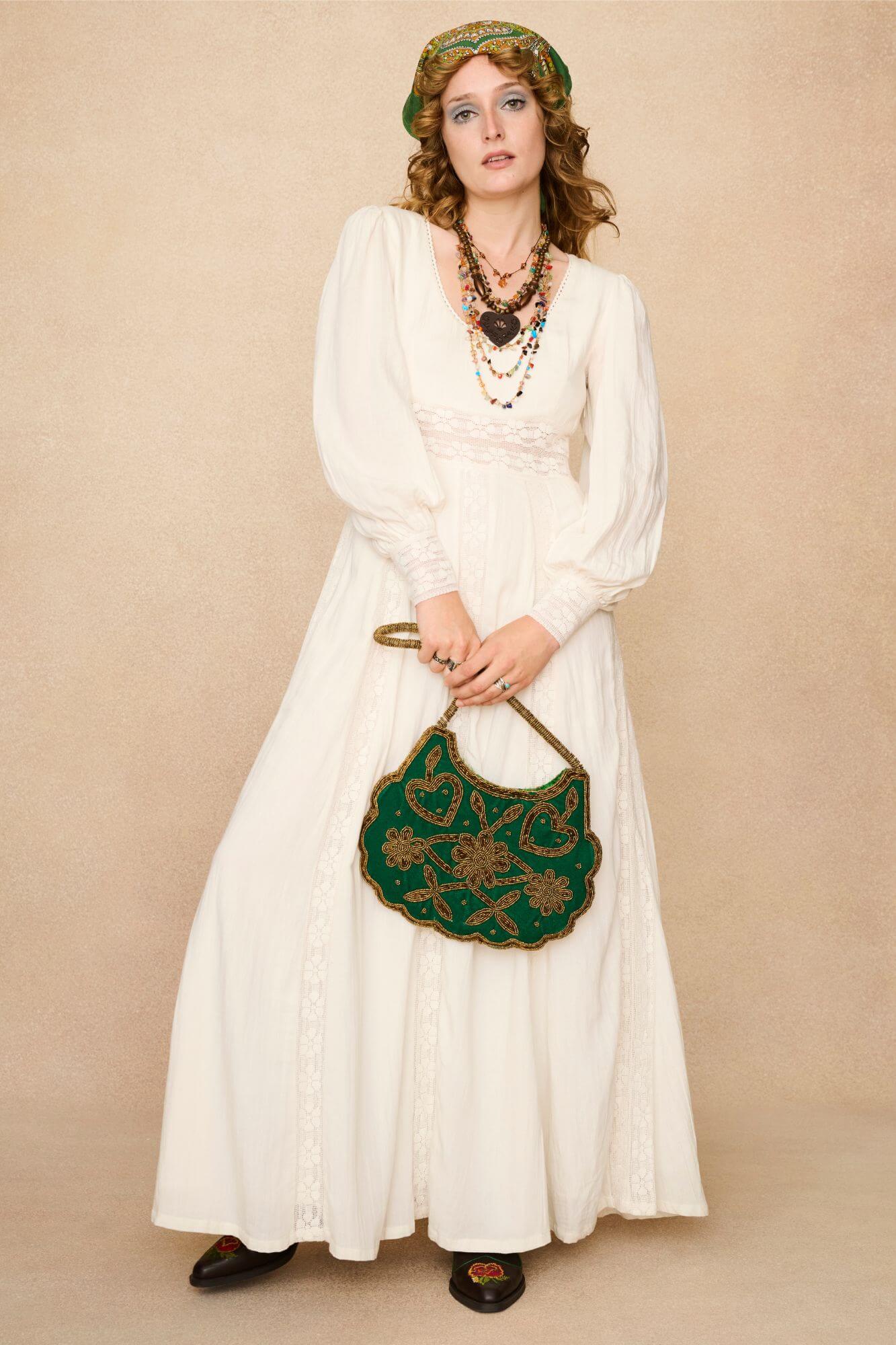 Woman in a long cream lace floaty 70s style maxi dress against a beige background - front view with bag