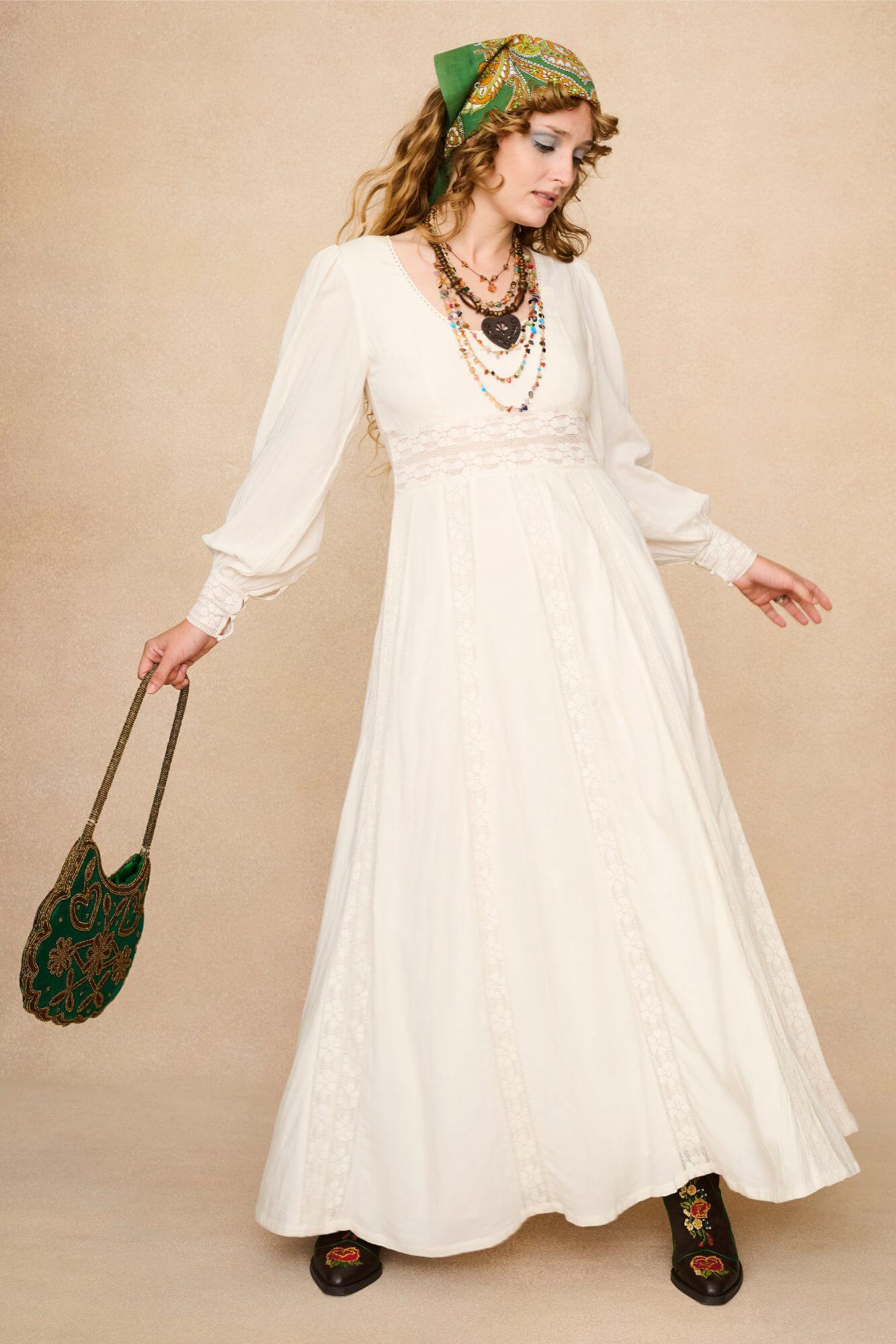 Woman in a long cream lace floaty 70s style maxi dress against a beige background - front view