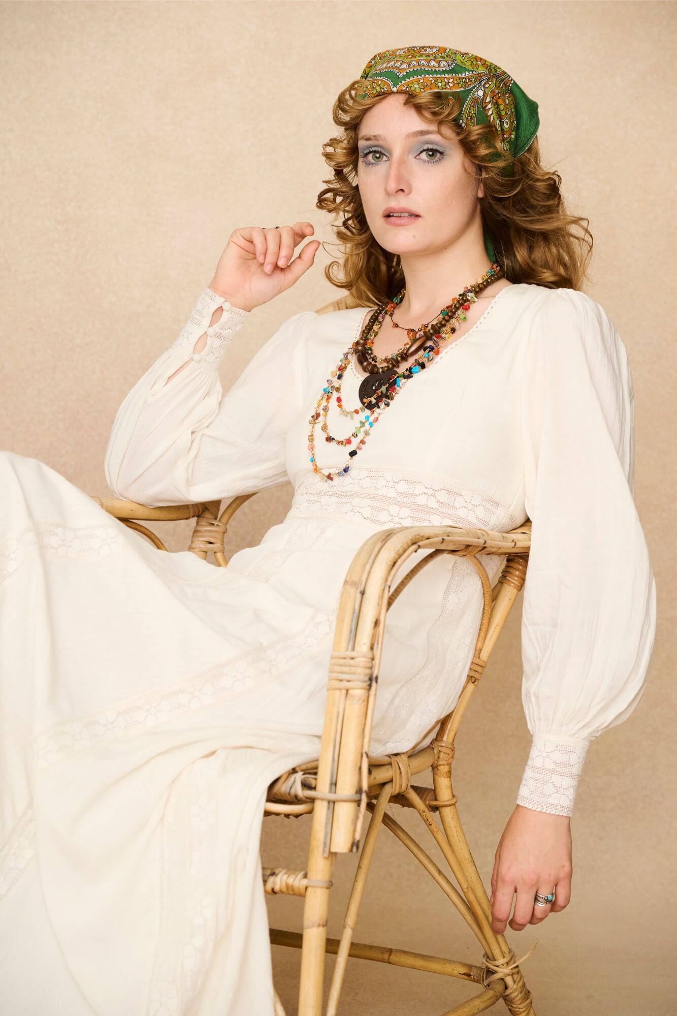 Woman in a long cream lace floaty 70s style maxi dress against a beige background - seated