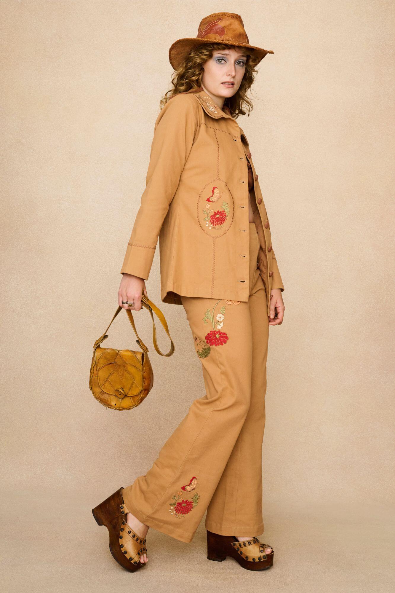 Model wearing a tan 70s style coat with embroidered floral patches against a beige background - outfit look