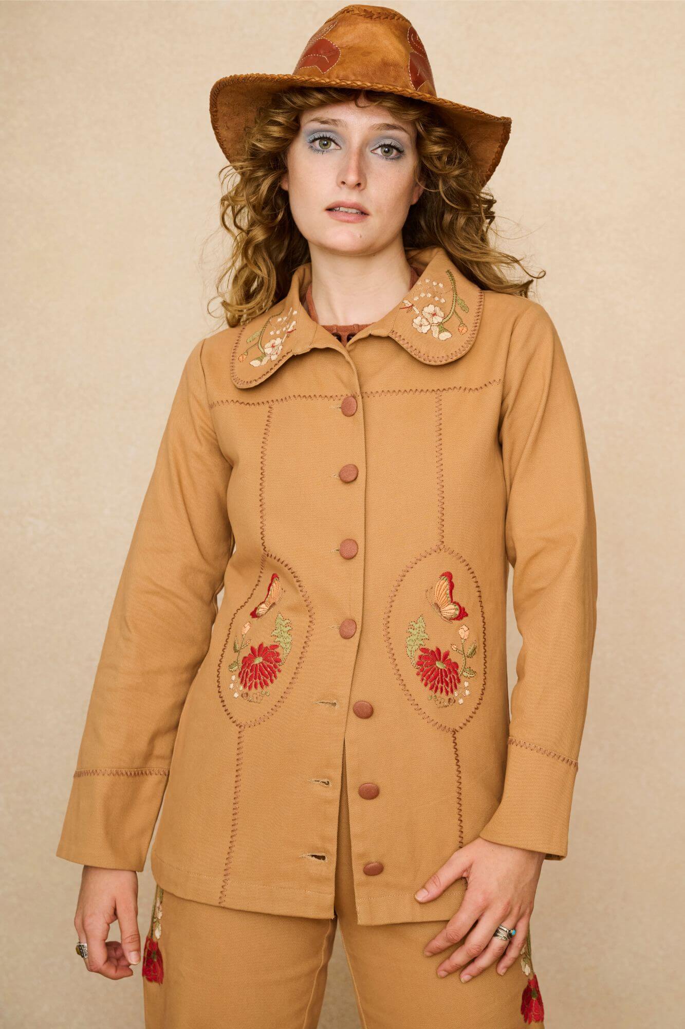 Model wearing a tan 70s style coat with embroidered floral patches against a beige background - close up