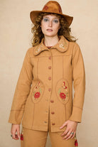 Model wearing a tan 70s style coat with embroidered floral patches against a beige background - close up
