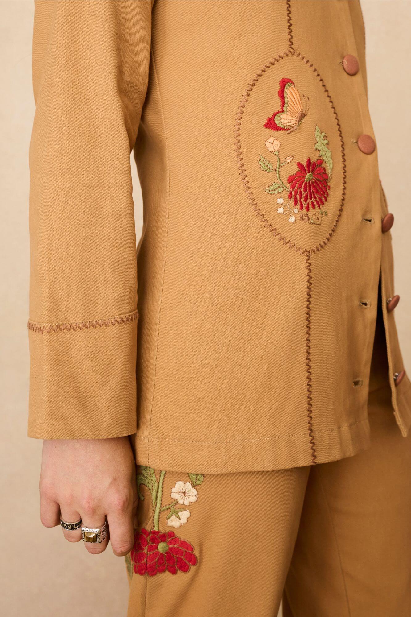 Model wearing a tan 70s style coat with embroidered floral patches against a beige background - close up of embroidery