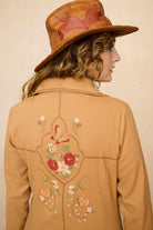 Model wearing a tan 70s style coat with embroidered floral patches against a beige background - close up of back embroidery