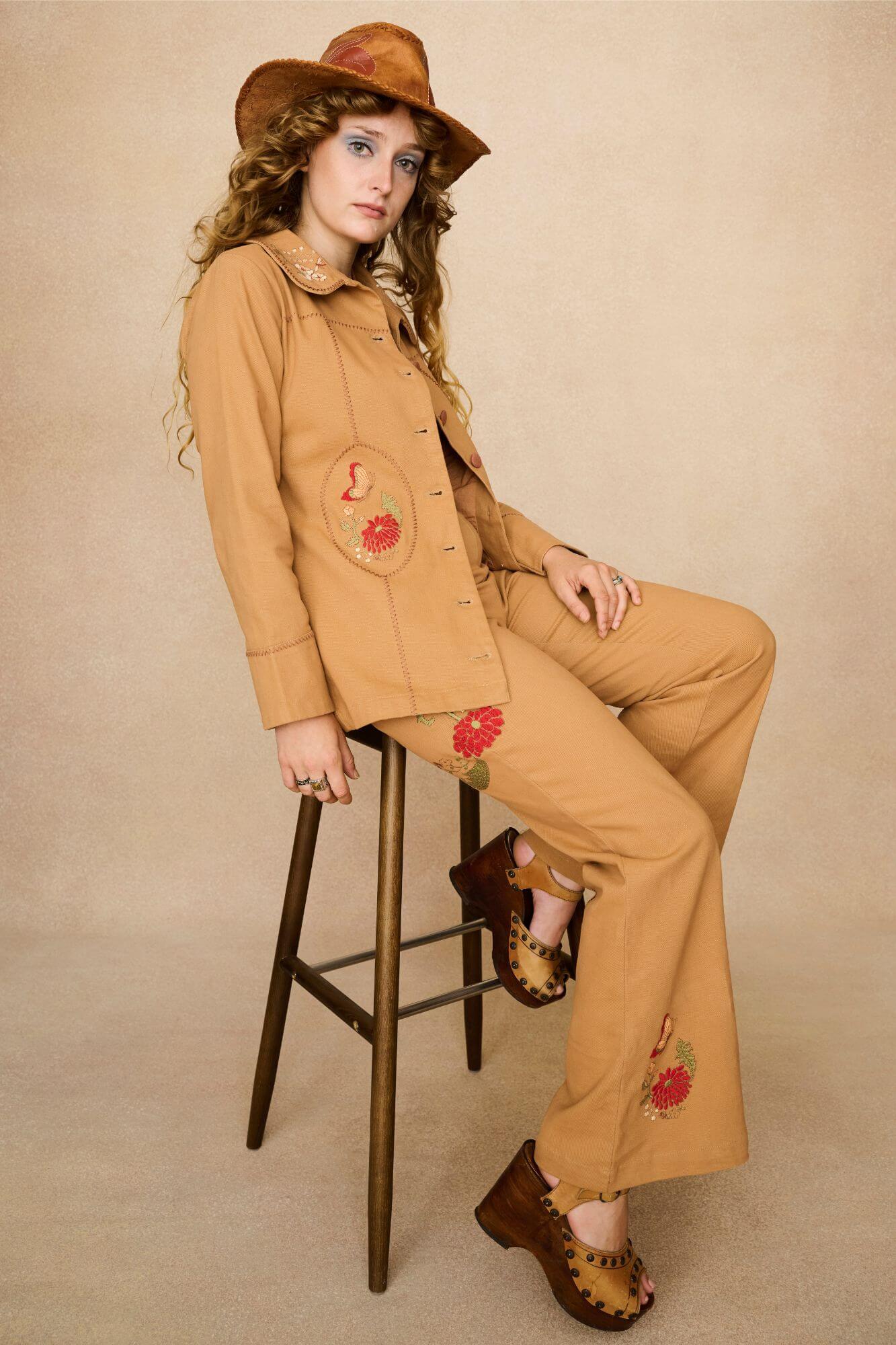 Tan-coloured 70s style trousers with floral embroidery on a beige background - seated view