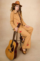 Tan-coloured 70s style trousers with floral embroidery on a beige background - model sitting on chair