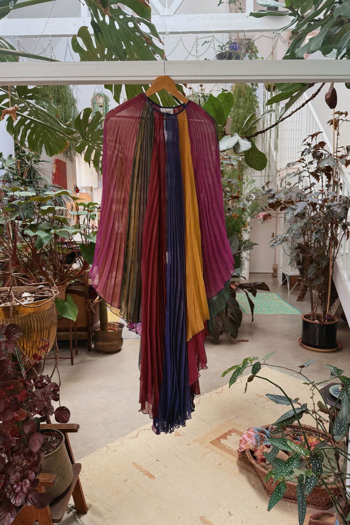 Colorful 70s cape dress hanging on a hanger in a greenhouse setting with plants and greenery.