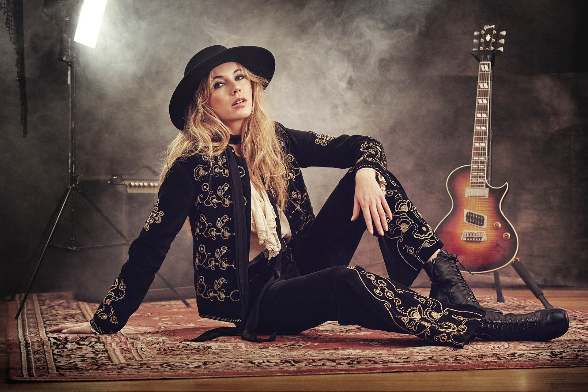 The Hippie Shake paint it black collection -model wears black velvet suit with gold embroidered embroidery sitting with a guitar on stage