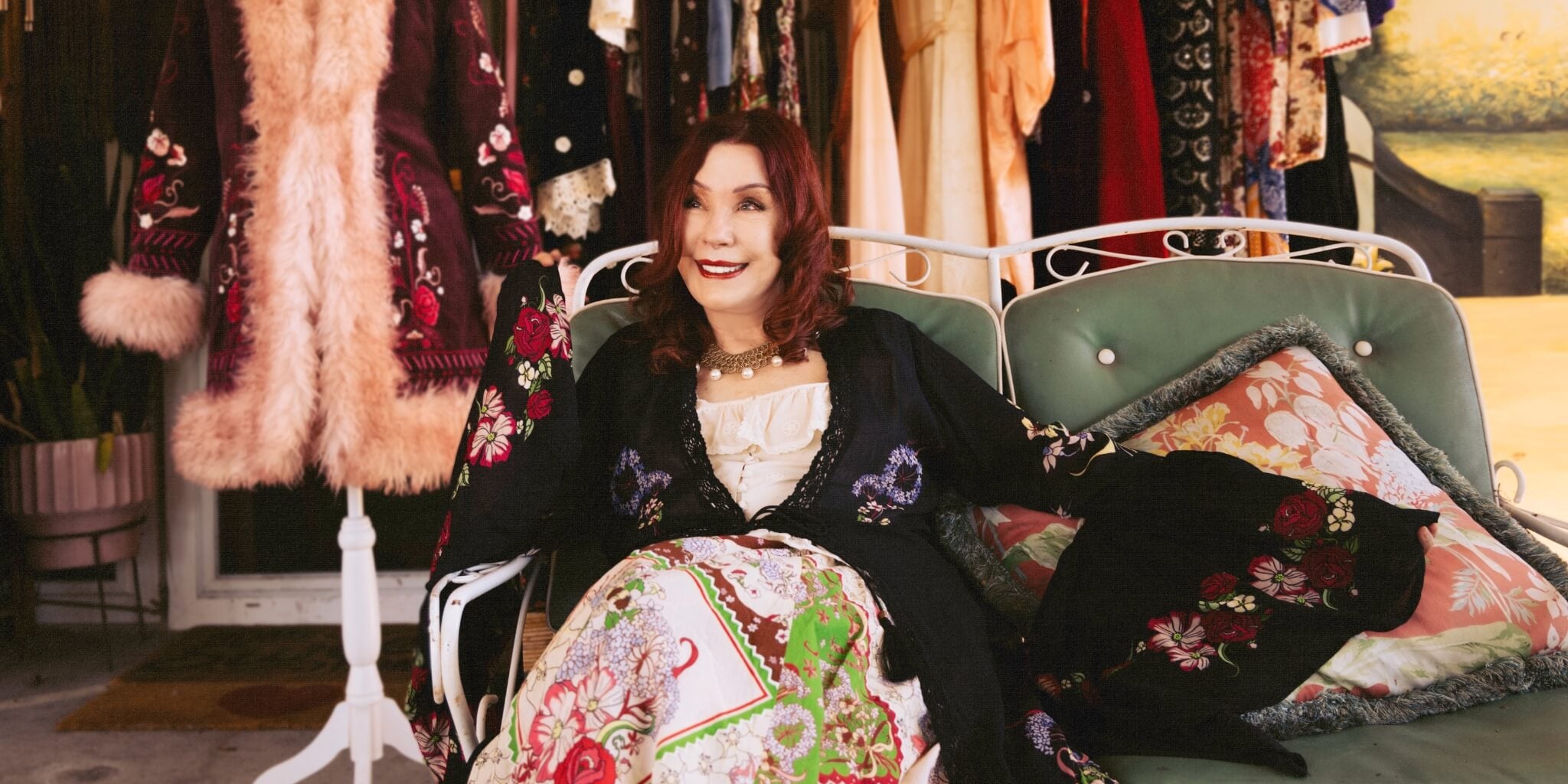 Pamela wearing black duster and handkerchief dress sitting in a room filled with colorful clothing