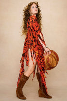 Model wearing a vibrant red and black patterned dress cape with fringe against a beige background - Side view