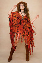 Model wearing a vibrant red and black patterned dress cape with fringe against a beige background - Front view with hat