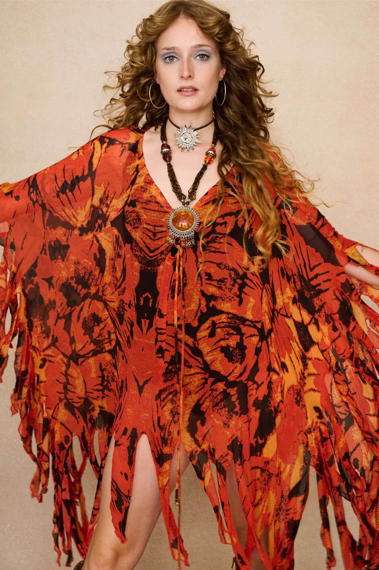 Model wearing a vibrant red and black patterned dress cape with fringe against a beige background - Close up view