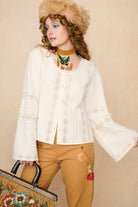 Woman wearing a white lace blouse and 70s style pants against a beige background - front view