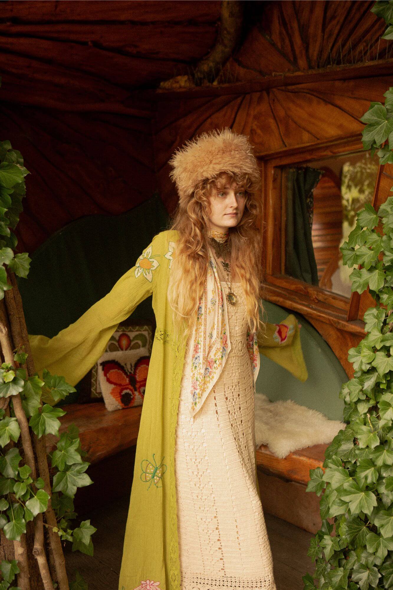 Woman in a green coat and fur hat standing in a rustic wooden cabin with greenery.