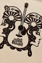 A close-up of a beige fabric surface printed with a black retro graphic featuring a guitar, stylized faces, stars, and the text 'the Hippie Shake'.