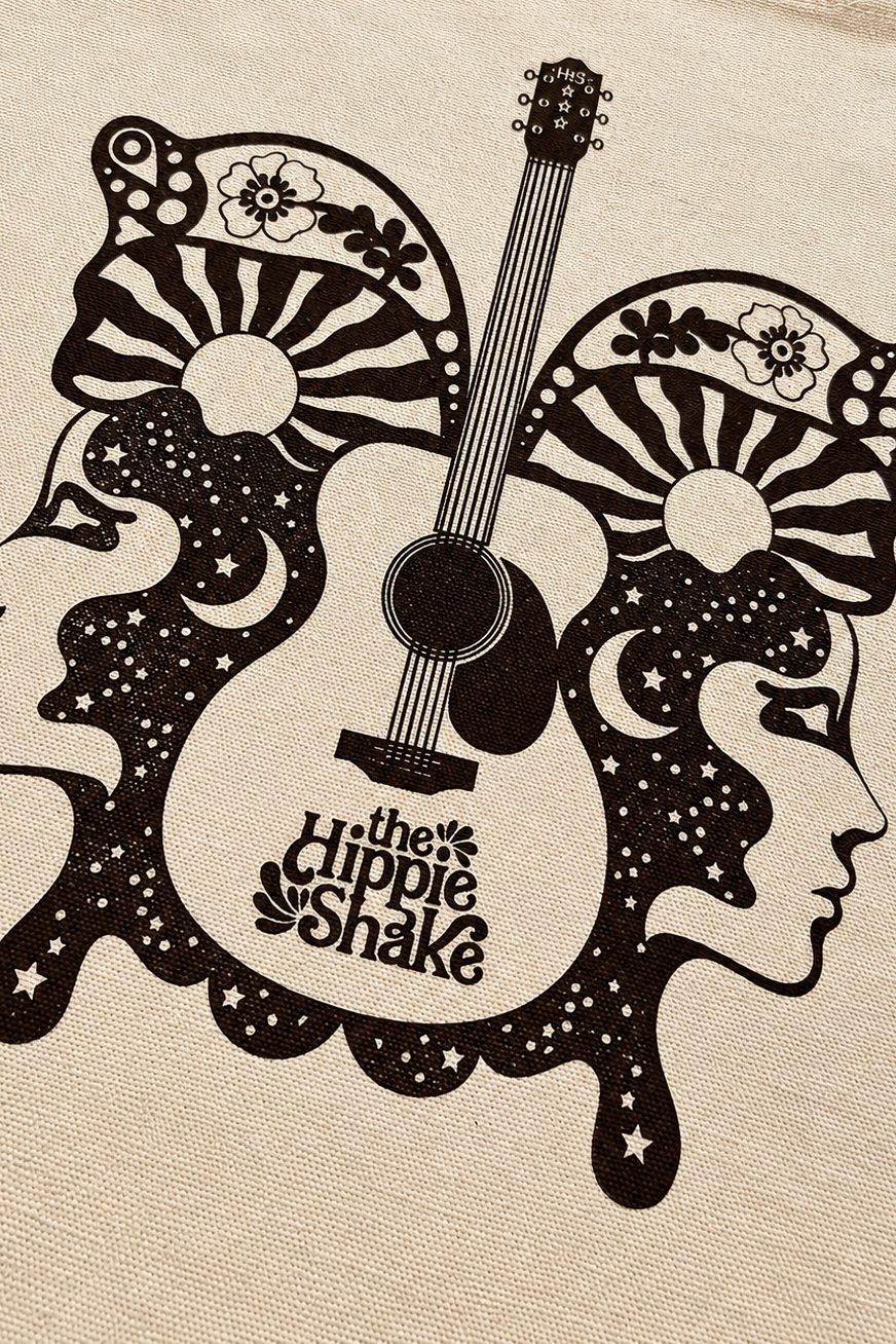 A close-up of a beige fabric surface printed with a black retro graphic featuring a guitar, stylized faces, stars, and the text 'the Hippie Shake'.